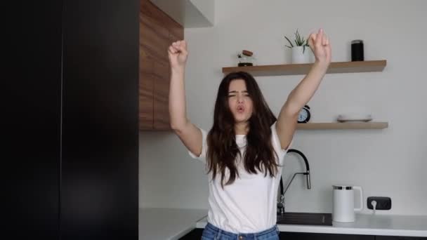 Mujer joven en la cocina durante la cuarentena. Disfrutando y gritando por la felicidad. Saludando con las manos y bailando. Chica alegre positivo emocionado . — Vídeos de Stock