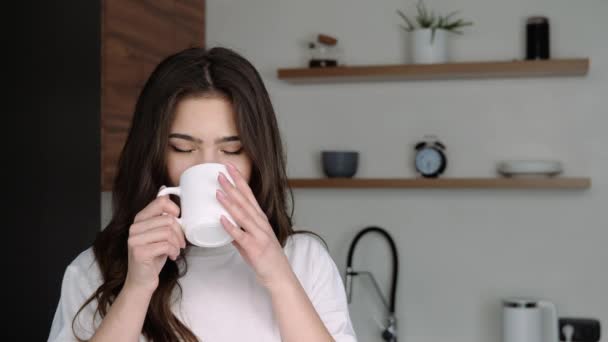 Mujer joven en la cocina durante la cuarentena. Hermosa bebida chica positiva de la taza blanca en cámara lenta. Posando en cámara con emociones alegres . — Vídeo de stock