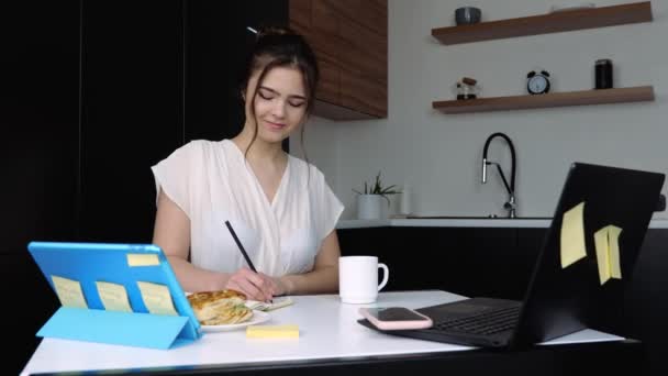 Jonge vrouw in de keuken tijdens quarantaine. Aan tafel zitten en studeren of leren. Werk op afstand met tablet, laptop en smartphone. Notities schrijven en graag thuis werken. — Stockvideo