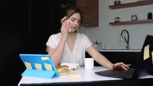 Jonge vrouw in de keuken tijdens quarantaine. Praten over de telefoon en werken aan de laptop. Thuis werken op afstand. Tafel met ontbijt op tafel. — Stockvideo