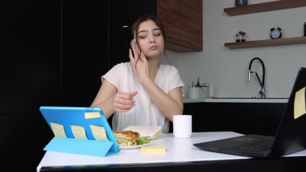 Mujer joven en la cocina durante la cuarentena. Chica sentarse en la mesa y tiene desayuno o almuerzo durante la conversación telefónica y descanso de trabajo . — Vídeo de stock