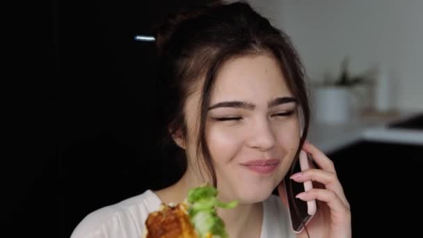 Mujer joven en la cocina durante la cuarentena. Chica hablando en el teléfono inteligente y comer delicioso sándwich sabroso. Desayuno en el almuerzo en la cocina . — Vídeo de stock