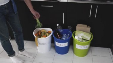 Karantina sırasında mutfakta genç bir adam. Plastik, kağıt ve organik atık için üç kova video. Sürtünme ve geri dönüşüm işlemi. Adam brokoliyi turuncu atık kovasına attı..
