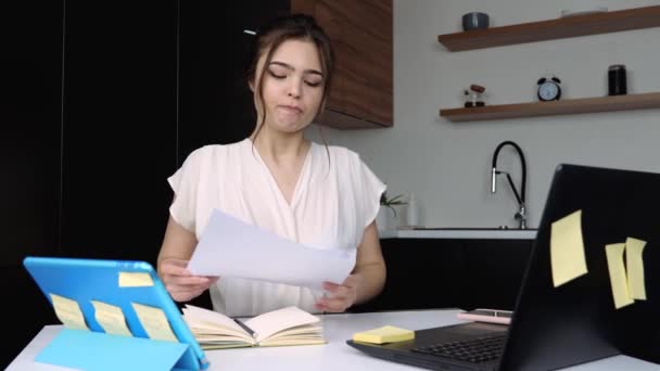 Mladá žena v kuchyni během karantény. Dívka píše na klávesnici notebooku a drží dokumenty v rukou. Volání na smartphone kvůli pracovním otázkám. — Stock video