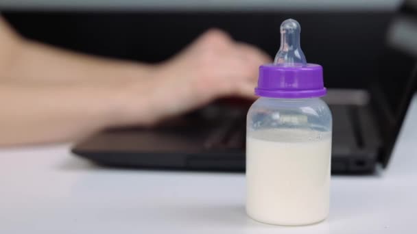 Young woman in kitchen during quarantine. Blurred background with female hands typing on laptop keyboard. Baby milk bottle in front. — Stock Video