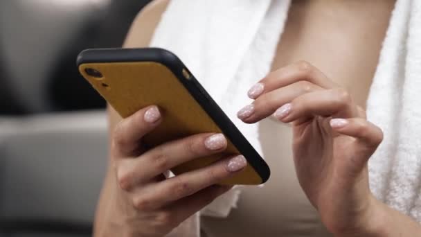 Una mujer joven hace ejercicio en casa durante la cuarentena. Primer plano de dedos femeninos escribiendo en la pantalla de teléfonos inteligentes. Charlando. Descanse después de hacer ejercicio. Toalla acostada en el hombro . — Vídeos de Stock