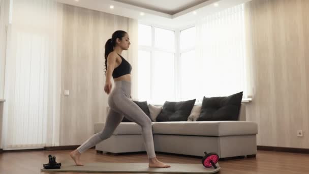 Una mujer joven hace ejercicio en casa durante la cuarentena. Párate derecho sobre una esterilla de yoga y toma una posición de asana guerrera en una sentadilla con las manos en alto. Hacer ejercicio y cuidar el cuerpo . — Vídeos de Stock