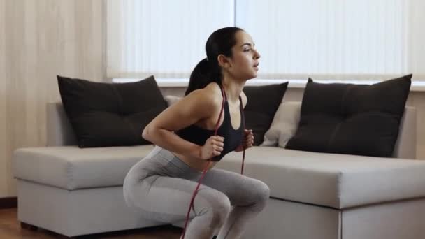 Una mujer joven hace ejercicio en casa durante la cuarentena. Hacer ejercicio en cuclillas con intensidad. Siéntate y ponte de pie varias veces. Formación en apartamento . — Vídeo de stock