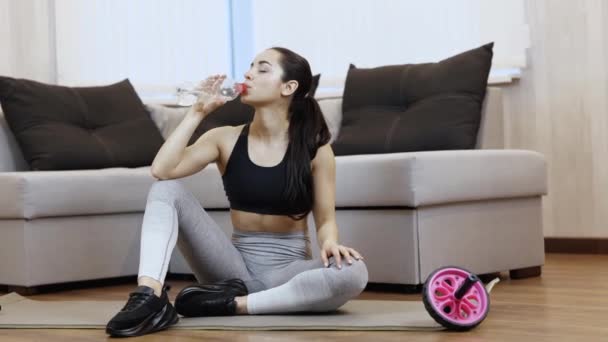 Ung kvinna har träning hemma under karantänen. Sitt på yogamattan och drick vatten efter träningen. Återfuktningstiden. Reklam. — Stockvideo