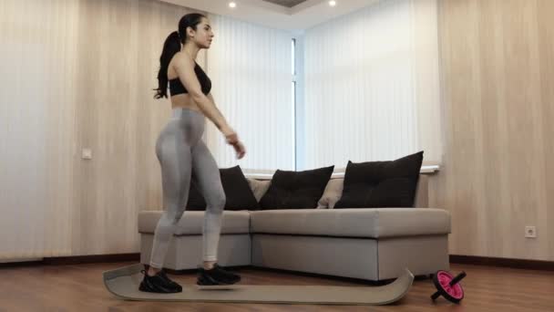 Mladá žena má doma během karantény cvičení. Přizpůsobivá štíhlá dívka dělá vždy jednu nohu dřepne. Aktivní cvičení v apartmánu. — Stock video