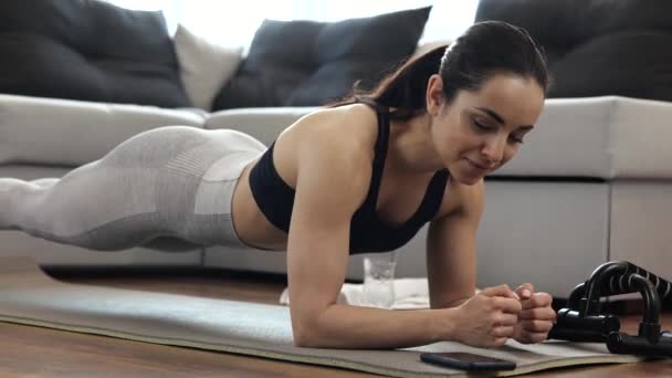 Jonge vrouw heeft thuis training tijdens quarantaine. Vrouwelijke persoon inschakelen stopwatch op smartphone en staan in plank positie. Gelukkig tevreden vrouw training thuis. — Stockvideo