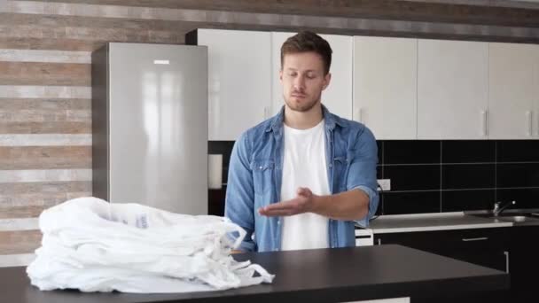 Jeune homme en cuisine pendant la quarantaine. Le gars pointe sur les sacs en plastique blancs et les nie. Afficher éco sac vert dans les mains et sourire. Achats responsables . — Video