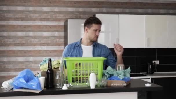 Un joven en la cocina durante la cuarentena. Tipo clasificando botellas de plástico y paquetes con bolsas. Ponlos en canasta verde. Uso responsable . — Vídeos de Stock