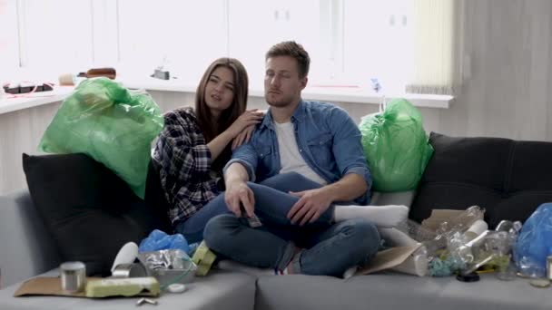 Jeune couple dans le salon pendant la quarantaine. Assis ensemble sur un canapé entouré de déchets recyclables. Regarder la télé et s'amuser. Vivre avec les ordures . — Video