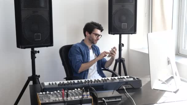 Jeune homme en studio d'enregistrement sonore. Asseyez-vous au clavier et utilisez un smartphone. Le type tapant sur l'écran du téléphone. La vie sur les réseaux sociaux en ligne. Utilisation d'une console ou d'un mixeur . — Video