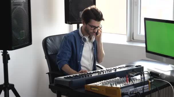 Joven en el estudio de grabación de sonido. Guy se sienta en el teclado y ajusta la consola de sonido antes de grabar. Hablando por teléfono. Pantalla digital verde . — Vídeos de Stock