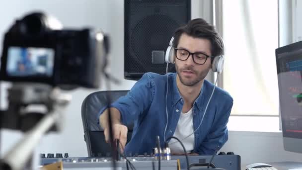 Jeune homme en studio d'enregistrement sonore. Blogueur vidéo expliquer à la caméra comment utiliser la console de mixage et de travailler avec elle. Écouter de la musique à travers des écouteurs . — Video