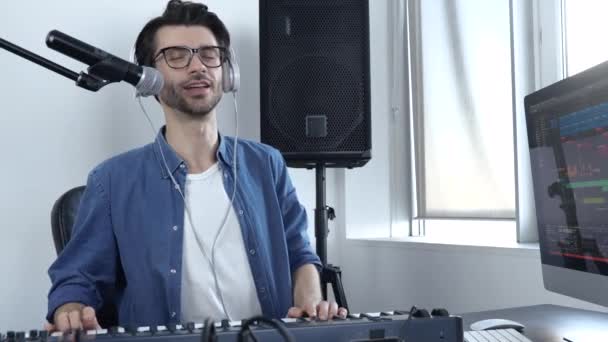 Mladý muž v nahrávacím studiu. Veselý pozitivní chlap hrající na klávesnici a zpívající. Neprofesionální hudebník se učí hrát. — Stock video