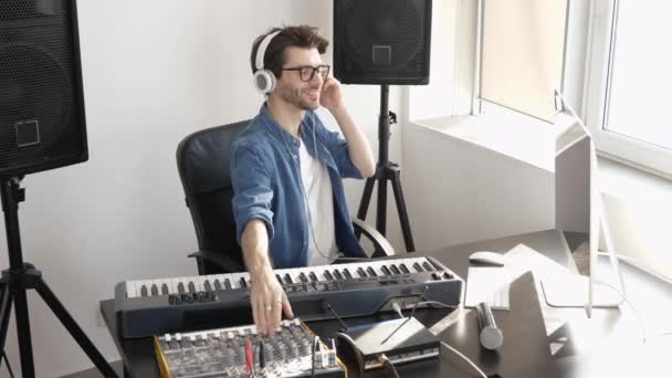 Молодой человек в студии звукозаписи. Сядьте за стол и используйте микшерную консоль. Слушать музыку через наушники . — стоковое видео