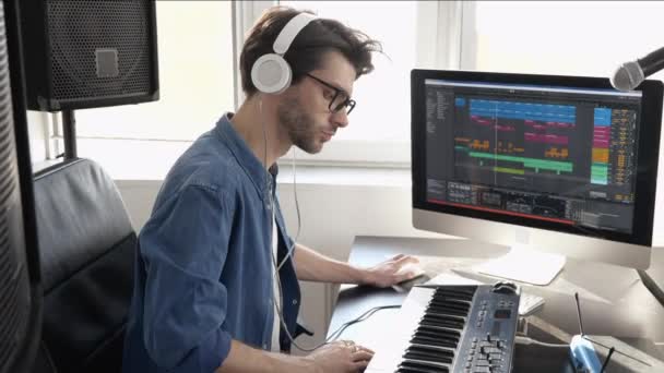 Mladý muž v nahrávacím studiu. Chlápek, co poslouchá hudbu přes sluchátka. Nahrávání hudby a přehrávání na klávesnici jako neprofesionální. — Stock video
