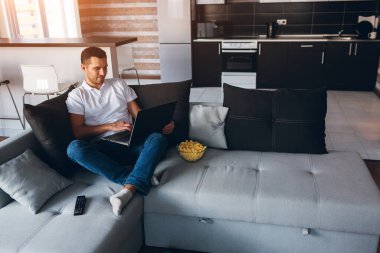 Genç adam kendi dairesinde televizyon izliyor. Mutlu olumlu erkek kişi dizüstü bilgisayarda çalışıyor. Uzaktan kumanda ofis dışında. Çalışkan insan eğlenir ve eğlenir. Odada tek başına.