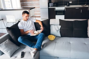 Genç adam kendi dairesinde televizyon izliyor. Sakin sakin konsantre adam couhc oturup film izledikten sonra kitap okuyor. Sessizce okumak. Çalışma ya da ofis dışında çalışma. Yetişkin öğrenci ya da okuyucu.
