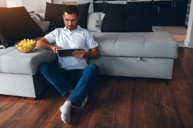 Genç adam kendi dairesinde televizyon izliyor. Film ya da program izliyordum. Yere otur ve tablet kullan. Uzaktan çalışma ya da ofis dışı. Evdeki iş adamı. Yetişkin eğitimi ya da çalışma.