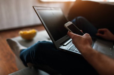 Genç adam kendi dairesinde televizyon izliyor. Görüntüyü kesin ve akıllı telefonu elinde tutan adamı kapatın. Dizüstü bilgisayarı bacaklarında. Film ya da program izle. Kendini eğlendir..