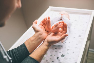Yeni doğmuş bir bebeğin ayaklarına yakın çekim. Genç baba pençelerinde bebek ayakları tutuyor. Küçük bebek çocuk masasında yatıyor. Pencerenin yanındaki bebek masasında yeni doğmuş bir çocuk var..