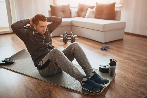 Joven hombre común ir a practicar deporte en casa. Estudiante de primer año se sienta en la alfombra y hace ejercicios abdominales. No es fácil empezar el entrenamiento solo en apatment. Principiante en acción. Equipamiento deportivo en el suelo . — Foto de Stock