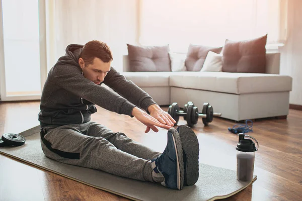 Ung vanlig man går in för sport hemma. Nybörjare eller amatör i träning aktivitet sitta på mattan och sträcka sig till tårna. Hårt arbetande kille tränar i apatment ensam. — Stockfoto