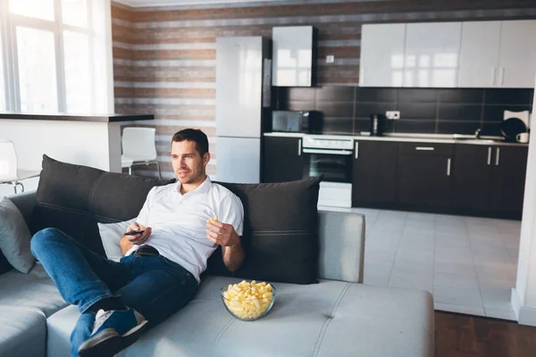 Young man watch tv in his own apartment. Sit alone on couch and eat snacks. Use remote control for switching tv channels. Looking for interesting movie or program.