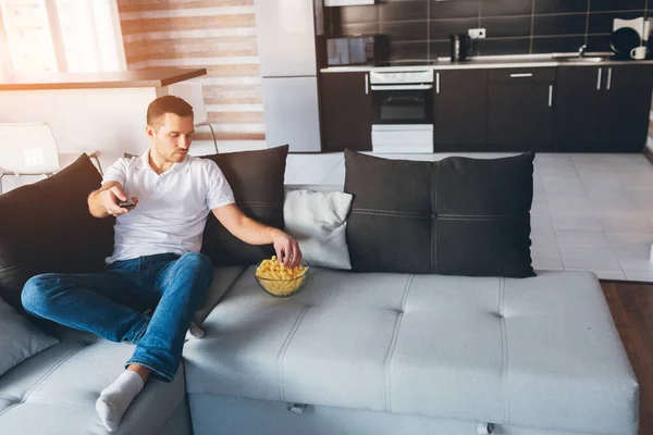 Young man watch tv in his own apartment. Serious concentrated guy hold remote control in hand and get snack from bowl. Watch movie or tv in room alone. Calm peaceful ordinary guy on picture.