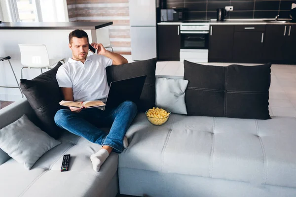 Young man watch tv in his own apartment. Serious concentrated guy reading book and talking on phone at same time. Hold opened laptop on knees. Studying or working. Remote out of office.