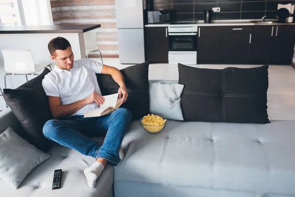 Un joven ve la televisión en su propio apartamento. Imagen de un tipo guapo e inteligente leyendo un libro solo en la habitación. Estudiar o leer para campesinos. Después de ver la película. Relájese y descanse . — Foto de Stock