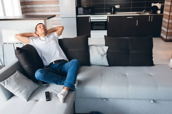 Young man watch tv in his own apartment. Guy enjoy his time in room. Sleeping joyful with hands under head. Lonely but happy. Rest and relax in new apartment. Daylight.