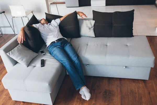 Young man watch tv in his own apartment. Picture of guy in deep sleep or daydream. Sitting or lying on couch and hold hands on black pillows. Sleep alone in room. Tired exhausted man.
