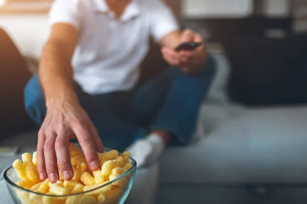 Young man watch tv in his own apartment. Cut view of guy reaching hand to bowl with unhealthy but tasty snacks for watching movie. Yellow and delicious.