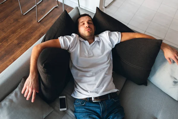 Young man watch tv in his own apartment. Sleeping tired and exhausted on couch alone. Daydreaming. Ordinary guy rest and relax. Hands lying on black pillows. Picture with up view.