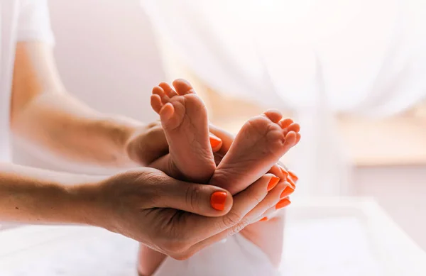 Close-up feet of a newborn baby. Young mother is holding baby feet in his hisands. Little infant is lying on the kids table. There is Newborn child on the baby changing table near window. — Stock Photo, Image