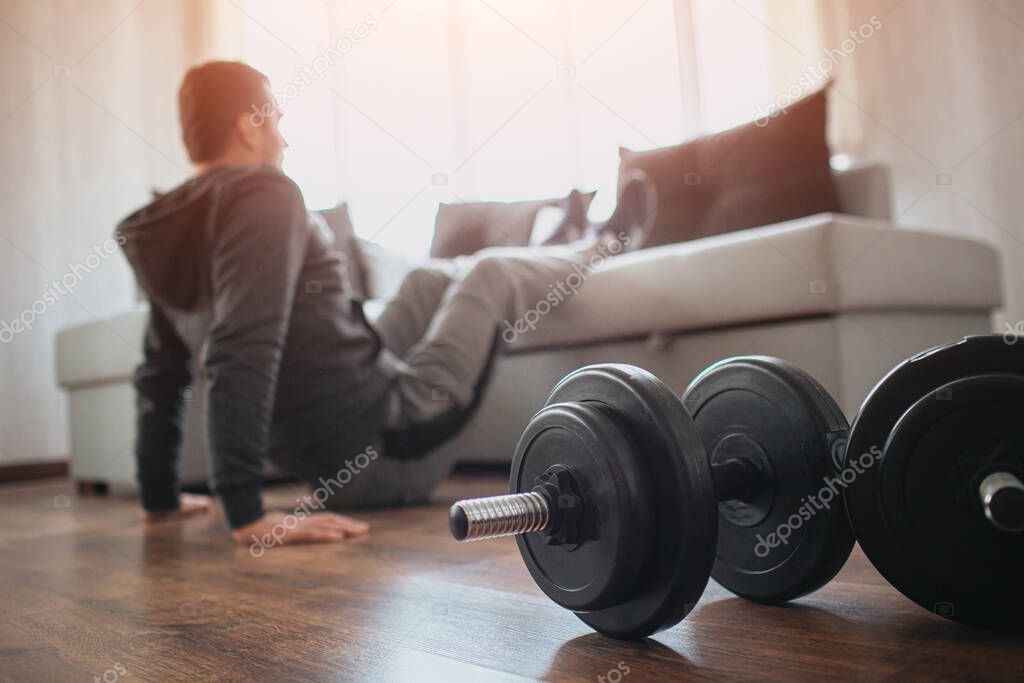 Young ordinary man go in for sport at home. Hardworking beginner sit on floor and hold legs on sofa. Freshmen trying to do abs exercising. Home alone doing workout. Dumbbells on floor.
