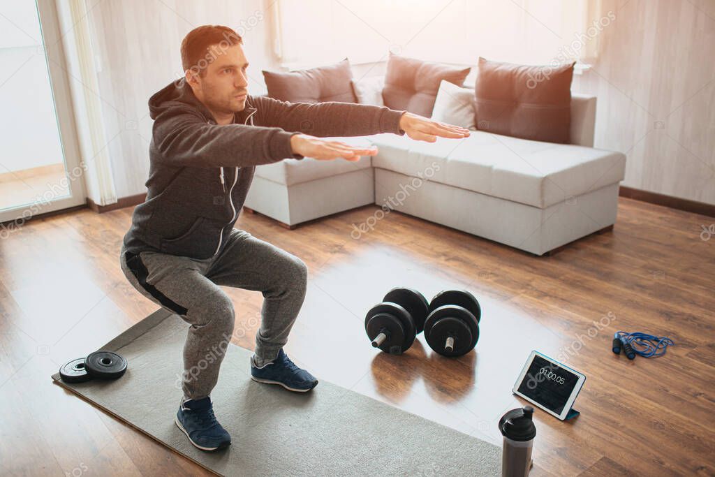 Young ordinary man go in for sport at home. Real picture of egular guy doing squats with stretch hands forward. Beginner or amateur has workout in apartment. Sport equipment on floor.