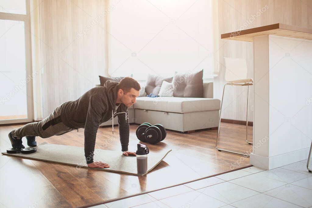 Young ordinary man go in for sport at home. Full size picture of regular ordinary guy stand in plank position alone in room. Beginner try to do his best and exercise. Hardworking real person.