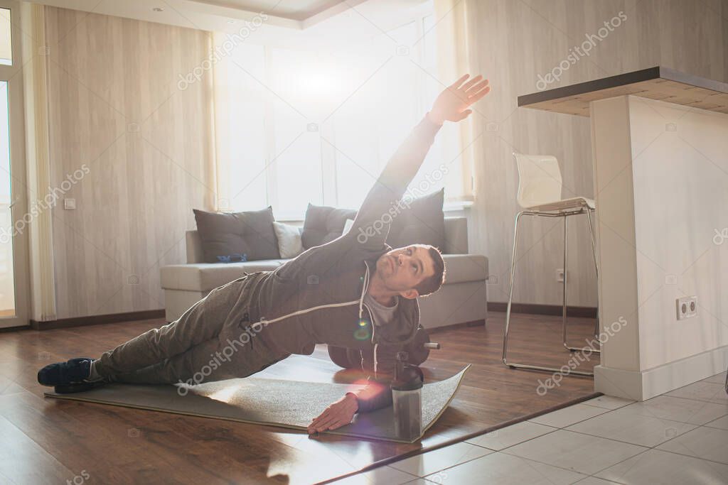 Young ordinary man go in for sport at home. Persistant freshman workout beginner stand in side plank position and look up on his stretched hand. Exercising alone in apartment. Ordinary guy on picture.