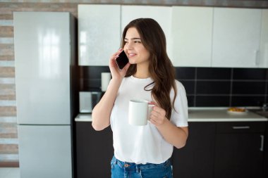 Karantina sırasında mutfakta genç bir kadın. Akıllı telefondan konuş ve gülümse. Karantina döneminde modern teknolojiler. Çevrimiçi çalışma.