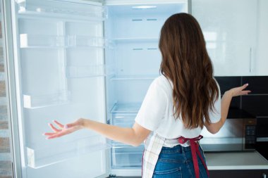 Karantina sırasında mutfakta genç bir kadın. İçinde yiyecek olmayan boş buzdolabı raflarına bak. Açım ve yemek pişiremiyorum. Kadının arkası, ne yapacağını bilemez..