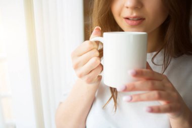 Karantina sırasında mutfakta genç bir kadın. Beyaz perdelerle pencerenin önünde dur ve fincanı çay ya da kahveyle tut. Yalnız içmek. Sakin sakin bir kadın. Görünümü kes.