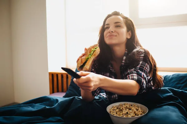 自宅で朝のベッドで若い美しい女性。テレビを見て興奮した幸せな女性の人。リモートコントロールを使用し、ポップコーンとサンドイッチを食べる。映画の時間. — ストック写真