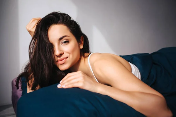 Jovem mulher bonita na cama da manhã em casa. Alegre positivo modelo feminino sexy posingo na câmera deitado e sorrir. Morena quente na foto no quarto ensolarado . — Fotografia de Stock