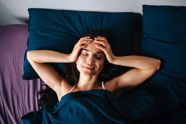 Молодая красивая женщина в утренней кровати дома. Спокойная, спокойная, расслабленная женщина-модель лежит одна. Держи руки за голову и улыбайся. Отдых и отдых
.